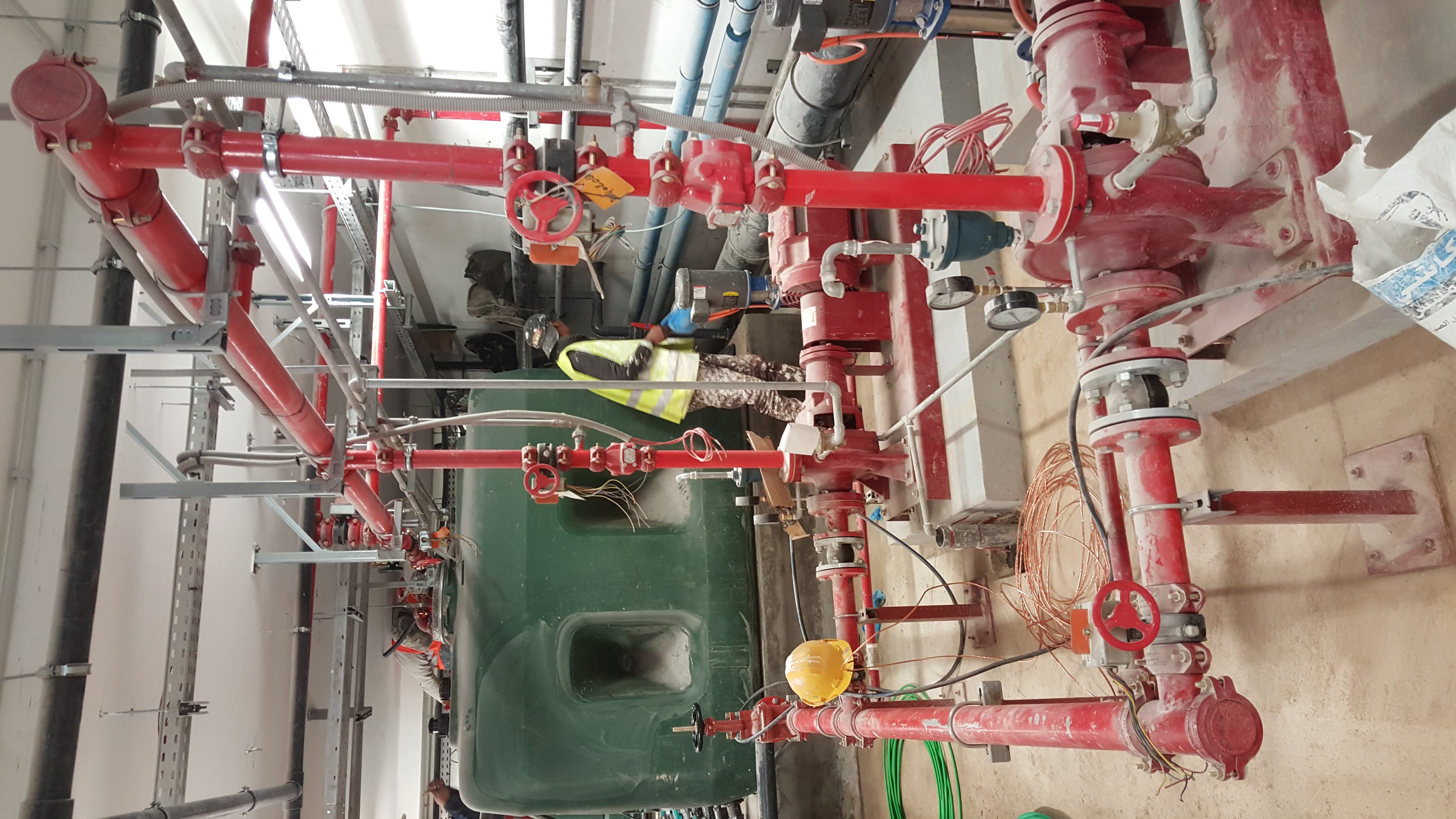 Système industriel de tuyauterie rouge avec vannes, réservoir vert et technicien en gilet haute visibilité.