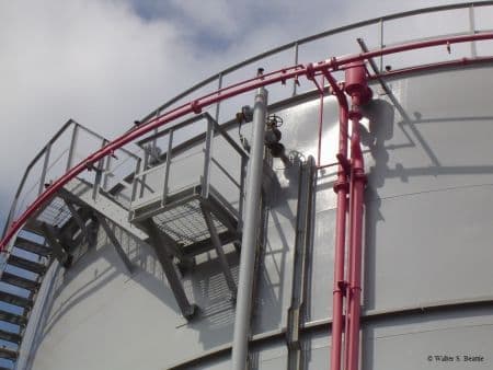 Grand réservoir industriel gris avec escaliers métalliques, plates-formes et tuyauterie rouge vif apparente.