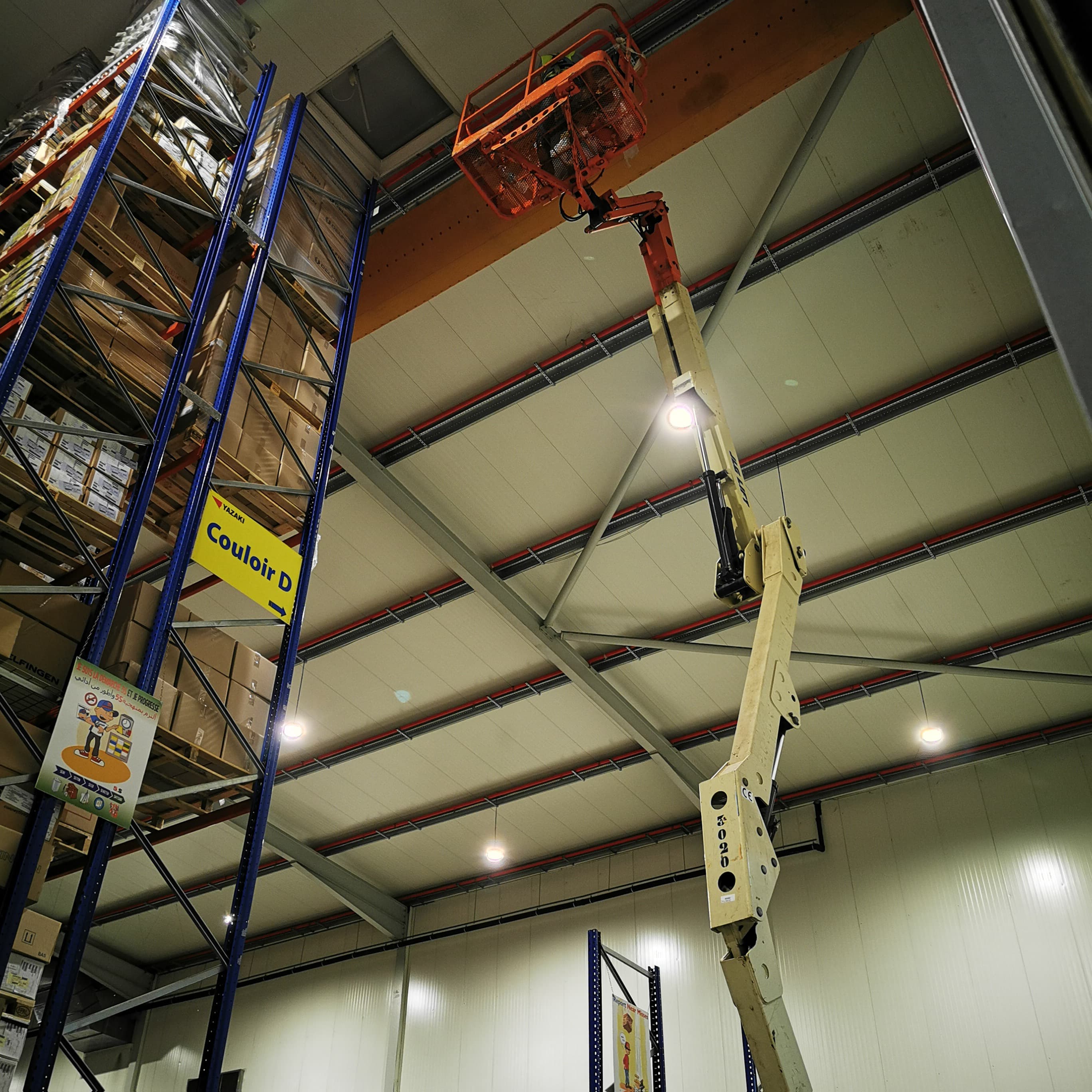 Bras articulé d'une nacelle élévatrice déployé vers le plafond d'un vaste entrepôt de stockage.