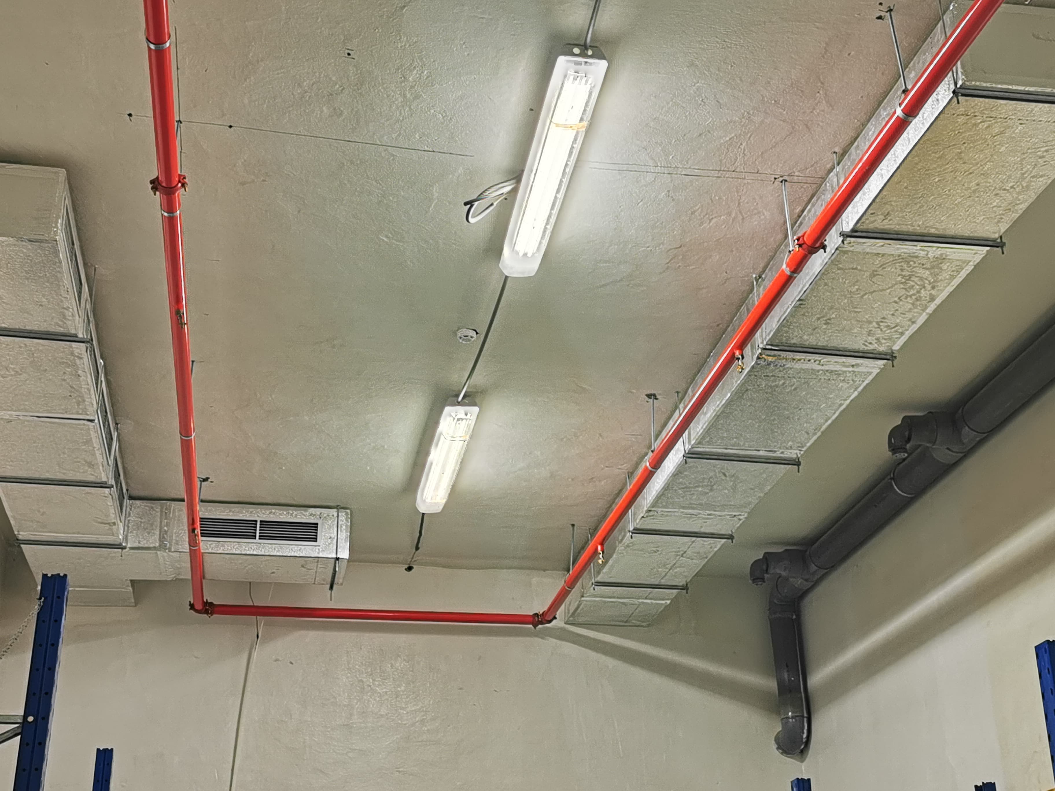 Plafond industriel avec conduits de ventilation, tuyaux rouges de protection incendie et éclairage fluorescent.
