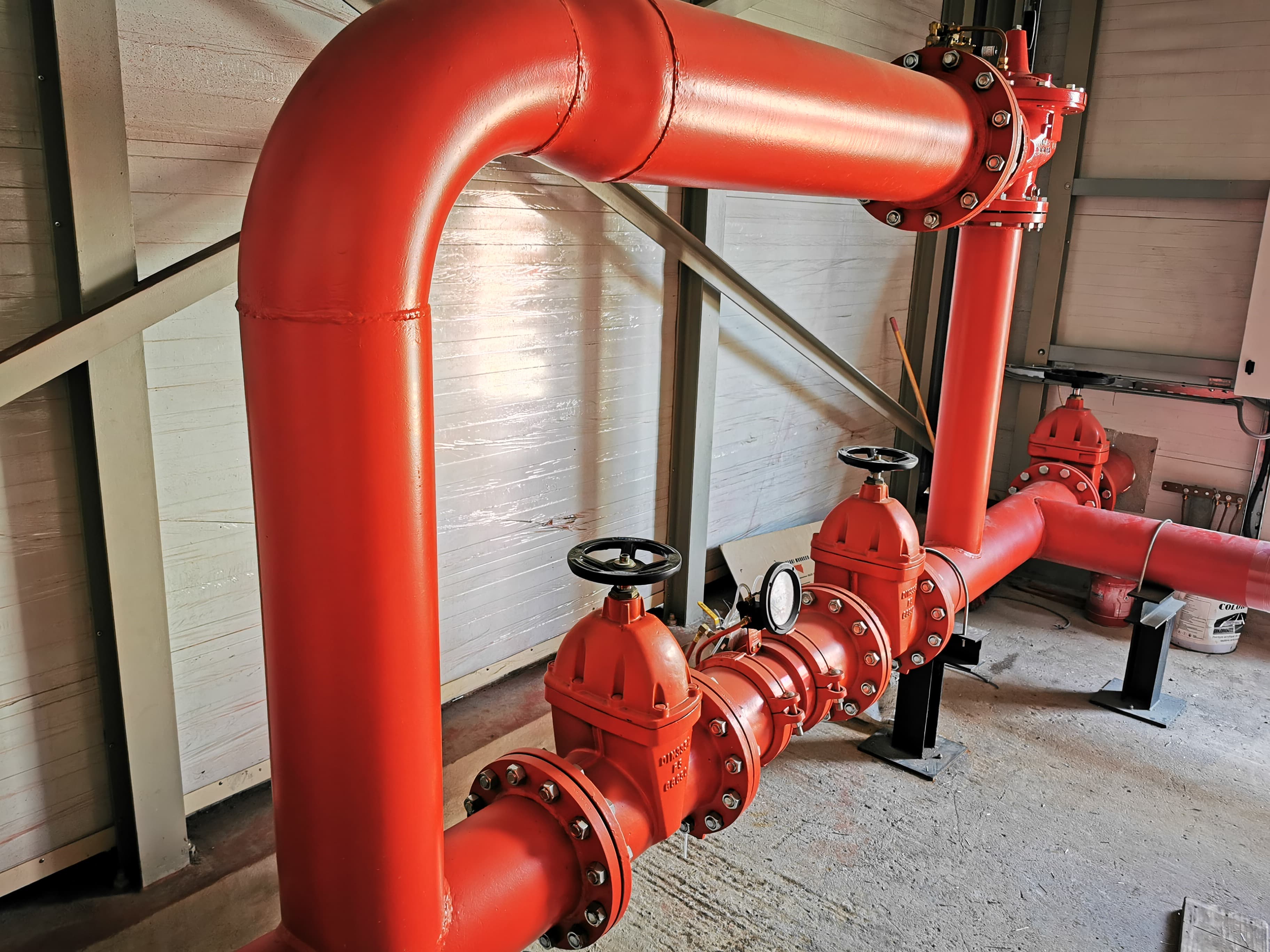 Grosses canalisations rouges avec vannes à volant noires et manomètre dans un bâtiment industriel.